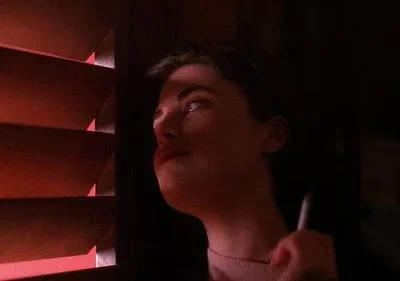 Jovem mulher em quarto escuro, rosto parcialmente iluminado por luz vermelha de persianas. Segura um cigarro, olhar pensativo. Clima de mistério.