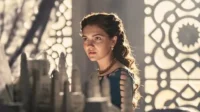 Mulher jovem séria com vestido verde-azulado e joias olha para modelos à frente. Luz quente ilumina seu cabelo e fundo ornamentado.