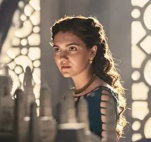 Mulher jovem séria com vestido verde-azulado e joias olha para modelos à frente. Luz quente ilumina seu cabelo e fundo ornamentado.