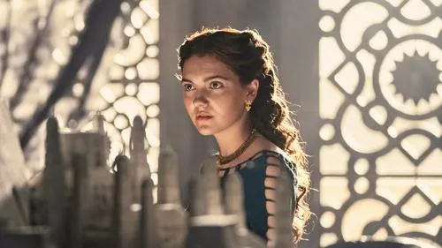 Mulher jovem séria com vestido verde-azulado e joias olha para modelos à frente. Luz quente ilumina seu cabelo e fundo ornamentado.