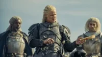 Três guerreiros medievais. Um loiro de armadura escura segura um pergaminho. Ao lado, um homem de dreads e um loiro, ambos armadurados. Clima épico.