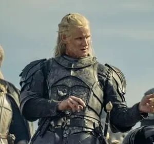 Três guerreiros medievais. Um loiro de armadura escura segura um pergaminho. Ao lado, um homem de dreads e um loiro, ambos armadurados. Clima épico.