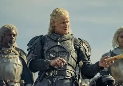 Três guerreiros medievais. Um loiro de armadura escura segura um pergaminho. Ao lado, um homem de dreads e um loiro, ambos armadurados. Clima épico.
