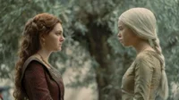 Duas mulheres jovens, uma de cabelo ruivo e outra loira platinada, se encaram tensas em um jardim. Vestes medievais.