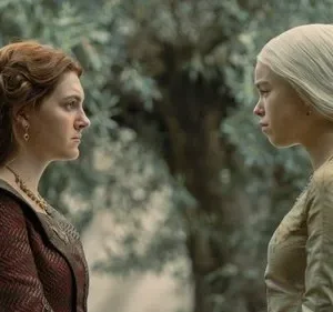 Duas mulheres jovens, uma de cabelo ruivo e outra loira platinada, se encaram tensas em um jardim. Vestes medievais.
