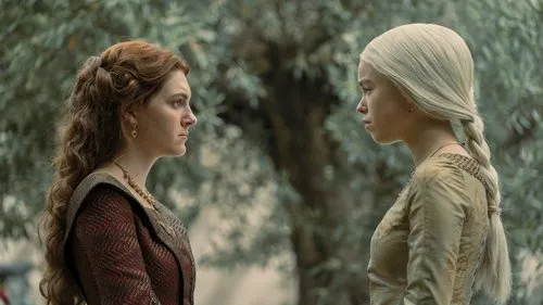 Duas mulheres jovens, uma de cabelo ruivo e outra loira platinada, se encaram tensas em um jardim. Vestes medievais.