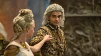 Homem de dreads prateados e casaco dourado ricamente bordado olha o espectador. Mulher loira em vestido claro segura seu braço. Cenário grandioso.