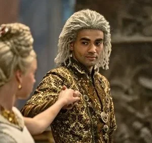 Homem de dreads prateados e casaco dourado ricamente bordado olha o espectador. Mulher loira em vestido claro segura seu braço. Cenário grandioso.