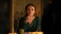 Jovem em vestido verde esmeralda e joias, com expressão séria, sentada à mesa em ambiente escuro e dramático.