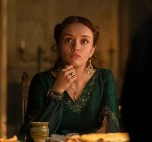 Jovem em vestido verde esmeralda e joias, com expressão séria, sentada à mesa em ambiente escuro e dramático.