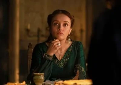 Jovem em vestido verde esmeralda e joias, com expressão séria, sentada à mesa em ambiente escuro e dramático.
