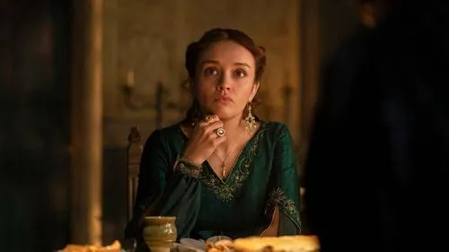 Jovem em vestido verde esmeralda e joias, com expressão séria, sentada à mesa em ambiente escuro e dramático.
