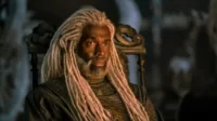 Homem com dreads brancos e barba, vestindo traje escuro e texturizado, sentado em um trono ornamentado. Ambiente escuro e sério.