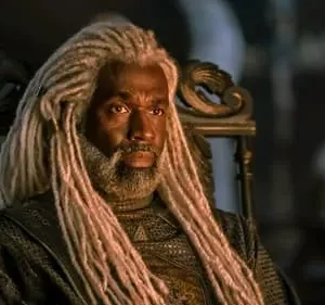 Homem com dreads brancos e barba, vestindo traje escuro e texturizado, sentado em um trono ornamentado. Ambiente escuro e sério.