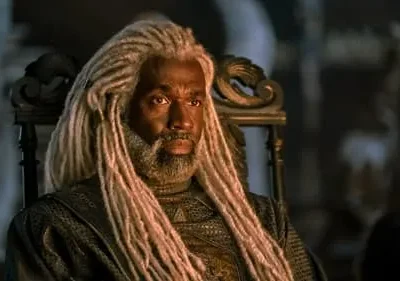 Homem com dreads brancos e barba, vestindo traje escuro e texturizado, sentado em um trono ornamentado. Ambiente escuro e sério.