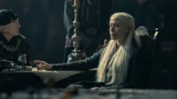 Mulher de cabelo loiro platinado e vestido escuro sentada à mesa, com expressão séria. Cena de época sombria, outras figuras ao fundo.