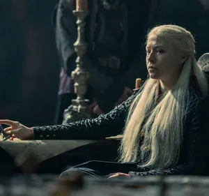 Mulher de cabelo loiro platinado e vestido escuro sentada à mesa, com expressão séria. Cena de época sombria, outras figuras ao fundo.