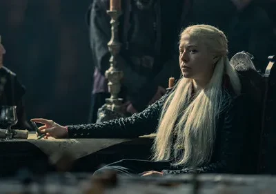 Mulher de cabelo loiro platinado e vestido escuro sentada à mesa, com expressão séria. Cena de época sombria, outras figuras ao fundo.