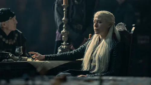 Mulher de cabelo loiro platinado e vestido escuro sentada à mesa, com expressão séria. Cena de época sombria, outras figuras ao fundo.