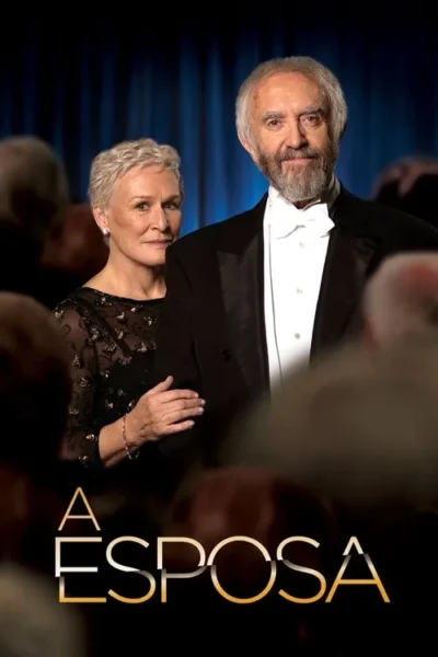 Homem idoso de smoking sorri, mulher de vestido preto e cabelo curto o observa com a mão no braço. Fundo de palco azul.