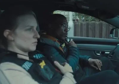 Dois policiais em um carro escuro. Mulher de colete à esquerda, homem de óculos no volante. Ambos sérios, com ar tenso e focado.