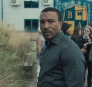 Homem sério olha para o espectador, com alunos apreensivos ao fundo de uma escola. Clima misterioso e tenso.