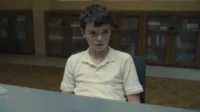 Jovem garoto de polo claro e expressão séria sentado à mesa. Estantes escuras com livros ao fundo. Clima sombrio.