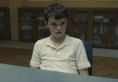 Jovem garoto de polo claro e expressão séria sentado à mesa. Estantes escuras com livros ao fundo. Clima sombrio.