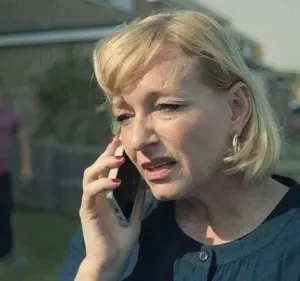 Em primeiro plano, mulher loira angustiada fala ao celular. Atrás, um homem embaçado parado no quintal. Clima tenso e misterioso.