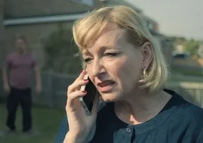 Em primeiro plano, mulher loira angustiada fala ao celular. Atrás, um homem embaçado parado no quintal. Clima tenso e misterioso.