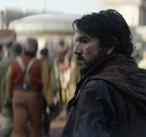 Homem barbudo de jaqueta escura em rua movimentada, olhando tenso para a esquerda. Fundo desfocado com figuras e edifícios.