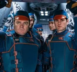 Dois homens sérios, de uniforme azul e laranja com bonés, seguram uma estrutura numa nave futurista. Outros tripulantes atrás.