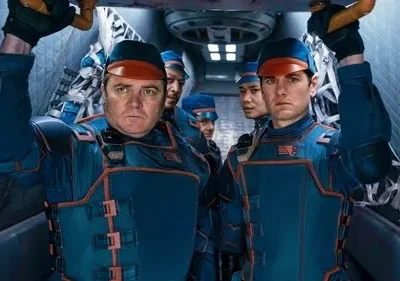 Dois homens sérios, de uniforme azul e laranja com bonés, seguram uma estrutura numa nave futurista. Outros tripulantes atrás.