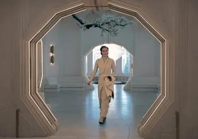 Mulher em traje futurista bege caminha por um arco octogonal iluminado. Cenário branco e moderno com galho florido e vista urbana.