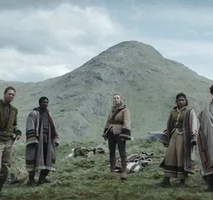 Seis figuras em vestes rústicas e camadas em paisagem montanhosa verde, sob céu cinzento. Sugere jornada ou desafio.