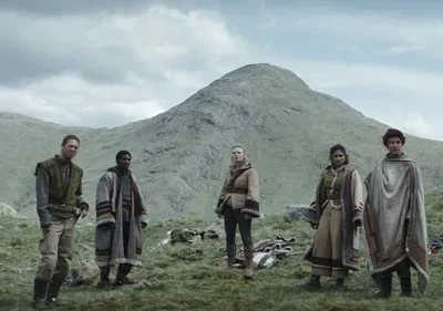 Seis figuras em vestes rústicas e camadas em paisagem montanhosa verde, sob céu cinzento. Sugere jornada ou desafio.