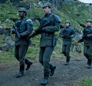 Quatro soldados em uniformes militares verde-cinza com rifles, marcham por uma trilha em terreno montanhoso e rochoso, sob um céu nublado.