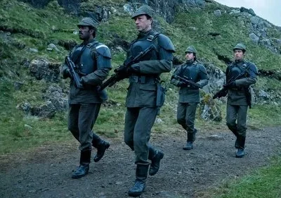 Quatro soldados em uniformes militares verde-cinza com rifles, marcham por uma trilha em terreno montanhoso e rochoso, sob um céu nublado.