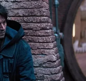 Um homem sério de jaqueta escura espreita atrás de um pilar. Ao fundo, duas figuras em armadura branca desfocadas patrulham. Clima tenso.