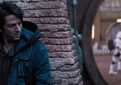 Um homem sério de jaqueta escura espreita atrás de um pilar. Ao fundo, duas figuras em armadura branca desfocadas patrulham. Clima tenso.