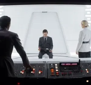 Oficial de costas opera painel. À frente, um homem sentado e uma mulher em uniforme branco observam em sala futurista e clara. Tensão.