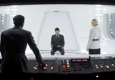 Oficial de costas opera painel. À frente, um homem sentado e uma mulher em uniforme branco observam em sala futurista e clara. Tensão.