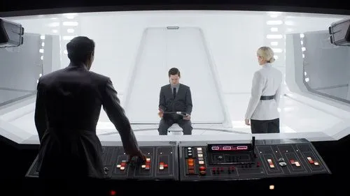 Andor – T01E08:Narkina 5 1 Oficial de costas opera painel. À frente,um homem sentado e uma mulher em uniforme branco observam em sala futurista e clara. Tensão.
