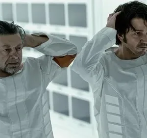 Dois homens, um mais velho e outro mais jovem, em uniformes brancos e laranja, com as mãos na cabeça, num ambiente estéril.