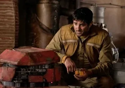 Homem pensativo de camisa marrom segura copos com líquido laranja, ao lado de um robô vermelho em ambiente industrial escuro.