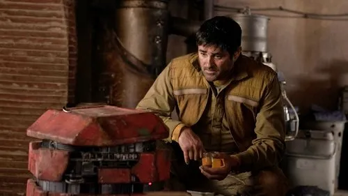Andor – T01E11:Filha de Ferrix 1 Homem pensativo de camisa marrom segura copos com líquido laranja,ao lado de um robô vermelho em ambiente industrial escuro.
