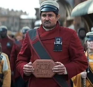 Líder sério de uniforme vermelho segura um bloco. Tropas em uniformes de cores diferentes o seguem em uma rua movimentada.