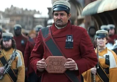 Líder sério de uniforme vermelho segura um bloco. Tropas em uniformes de cores diferentes o seguem em uma rua movimentada.