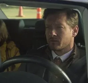 Homem sério dirige um carro, menina apreensiva de jaqueta amarela está no banco de trás. Atmosfera tensa.