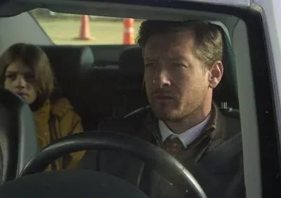 Homem sério dirige um carro, menina apreensiva de jaqueta amarela está no banco de trás. Atmosfera tensa.